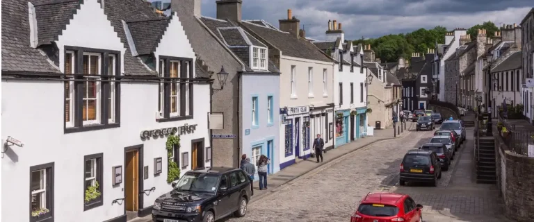 South Queensferry High Street with colourful historic buildings, cobbled paving, and parked cars, viewed on a bright day with the hill rising behind the town.