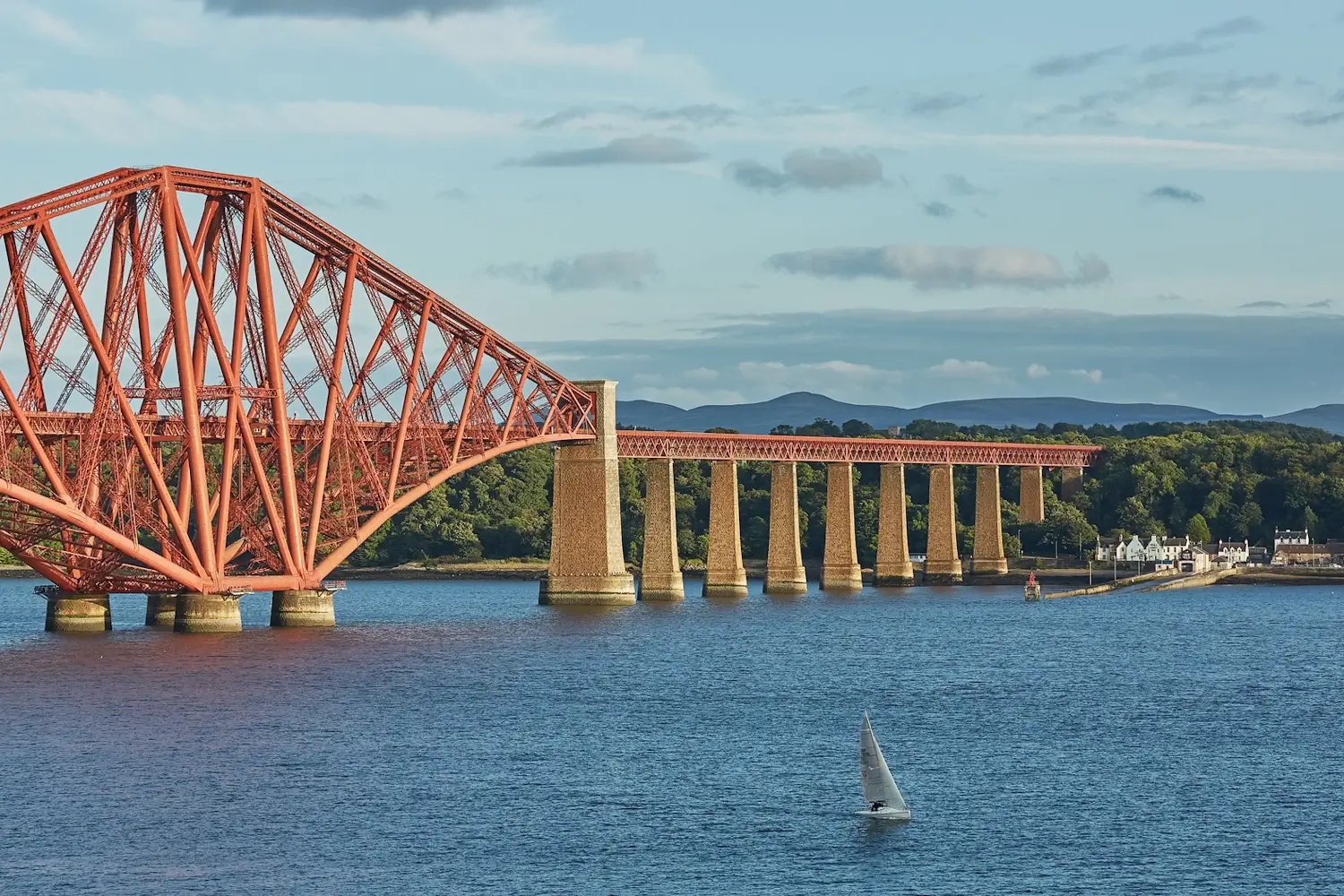 The Forth Rail Bridge stretching over the Firth of Forth near South Queensferry Cruise Port, with a small sailboat on the water.