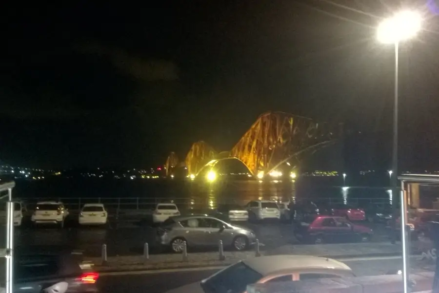 Night view of the Forth Bridge from the Railbridge Bistro, with cars parked along the waterfront below.