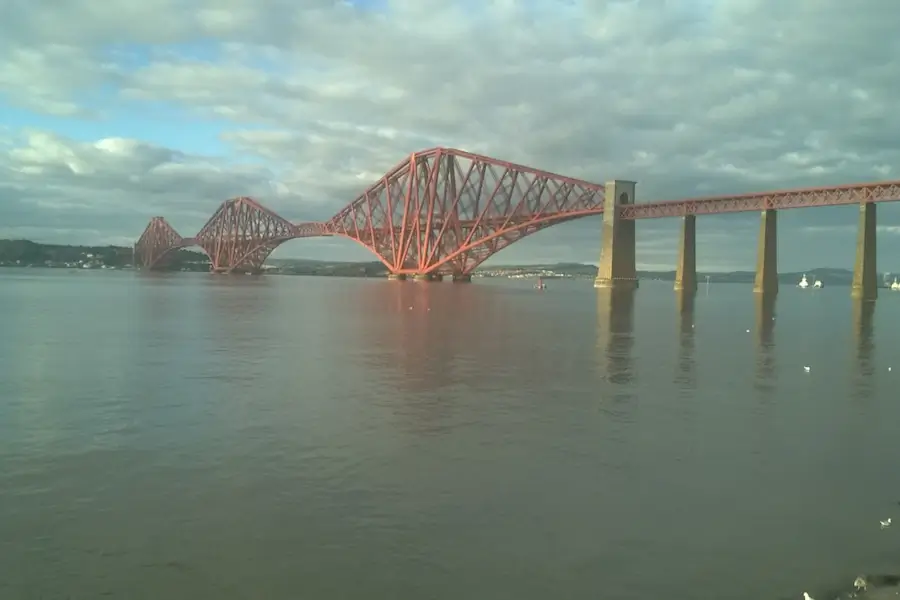 Forth Rail Bridge photographed from the shoreline in South Queensferry, with calm water reflecting the red steel spans and stone piers under a cloudy sky.