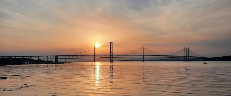 Sun setting behind the Forth bridges in South Queensferry, with calm water reflecting the light and bridge towers across the Firth of Forth.