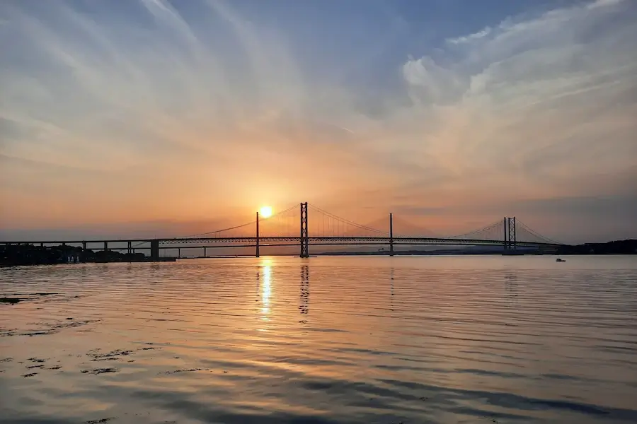 Sunset behind the Forth Road Bridge in South Queensferry, with calm water reflecting the bridge towers and orange evening sky across the Firth of Forth.