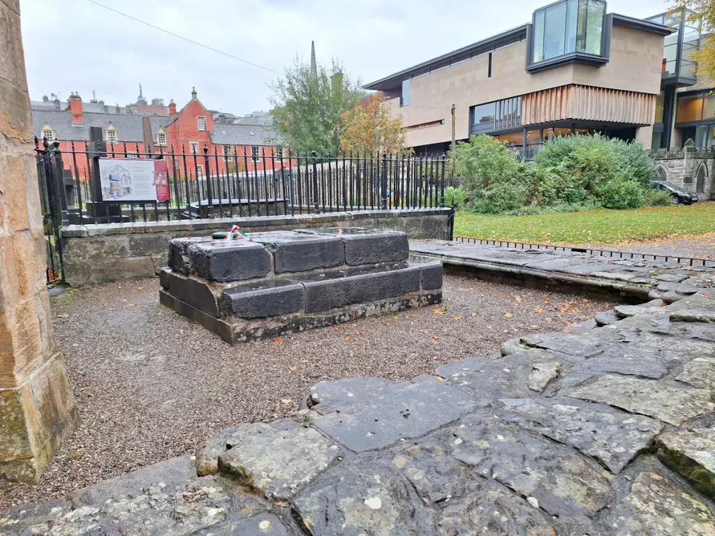 Remains of St Margaret’s shrine in the grounds of Dunfermline Abbey, railed off, with modern buildings behind